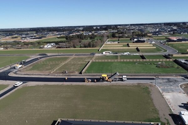 Subdivision roading and stormwater systems installation with a construction contractor laying road base layers, demonstrating civil construction and water reticulation infrastructure development for new housing areas.