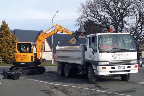 An excavator loads spoil into an On Grade dump truck during street excavation works, part of drainage and water reticulation upgrades in Canterbury.
