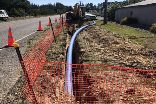 Open trench along a rural roadside with a newly installed blue watermain pipe, safety cones and fencing, and an excavator working on the water supply upgrade.