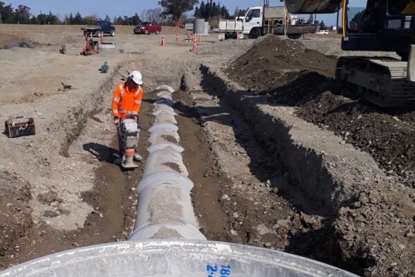 Excavator and crew installing large stormwater culvert sections in a long trench, preparing drainage infrastructure for a commercial development.