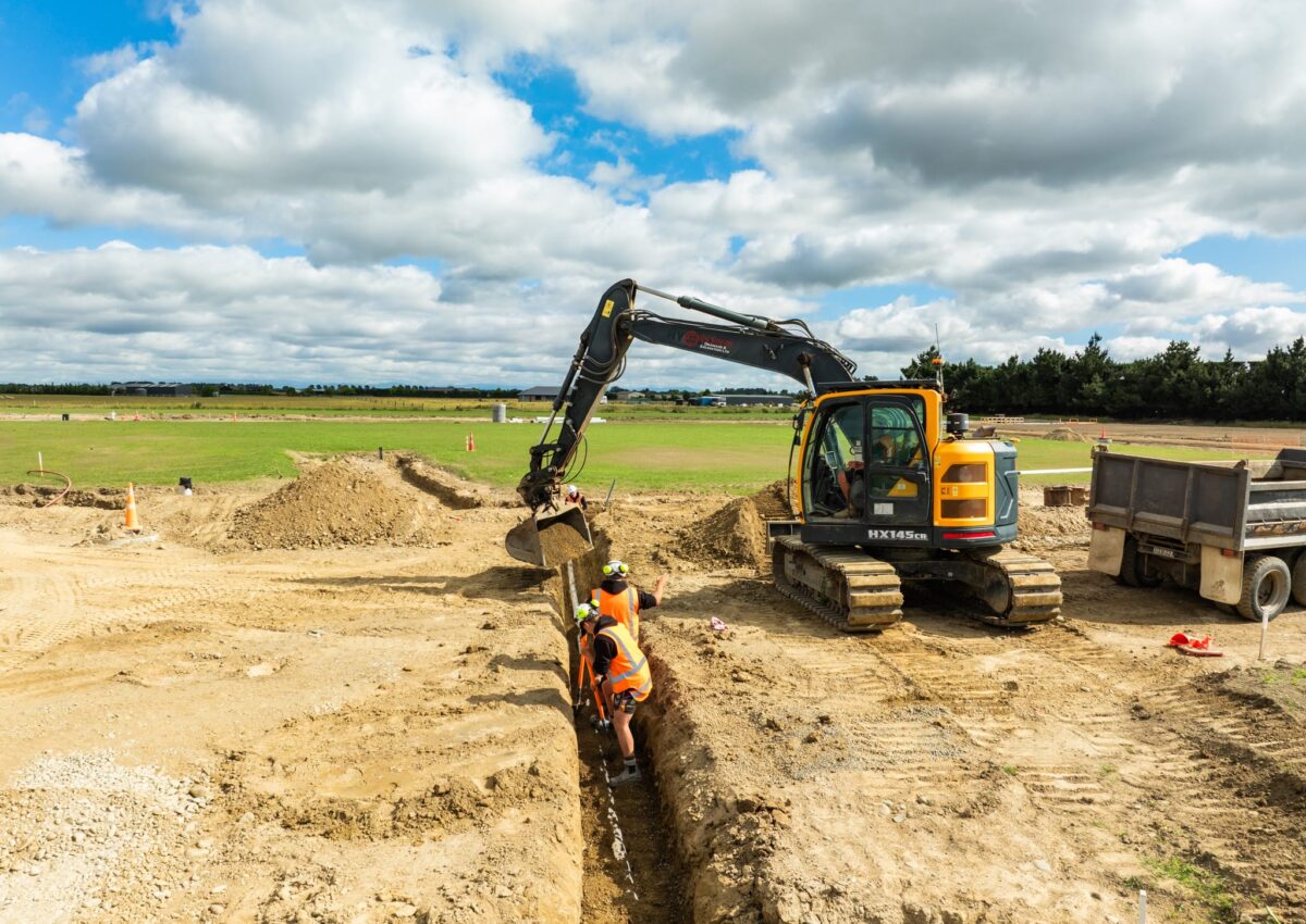 Excavator assisting civil contractors installing storm water drainage and water reticulation pipes in a deep trench during subdivision earthworks, supporting wastewater reticulation systems and civil construction works on a Christchurch development site.