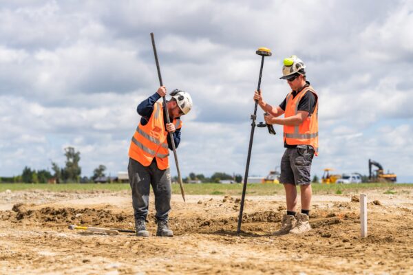 Two workers marking out levels on a civil construction site, carrying out surveying, storm water drainage planning, wastewater treatment system preparation and land excavation for subdivision earthworks.