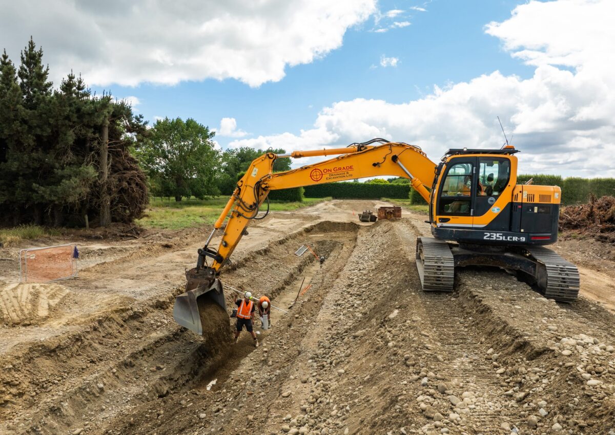 Excavator assisting workers inside a deep trench as civil contractors complete storm water drainage system installation, wastewater reticulation, sanitary sewer pump station preparation and subdivision earthworks for a major construction project.